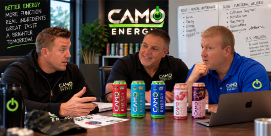 CAMO Energy founders Matthew Mallory, Adam Mallory and Casey Hill in conversation around a boardroom table lined with cans of CAMO Energy, with a CAMO Energy sign on the wall behind them.